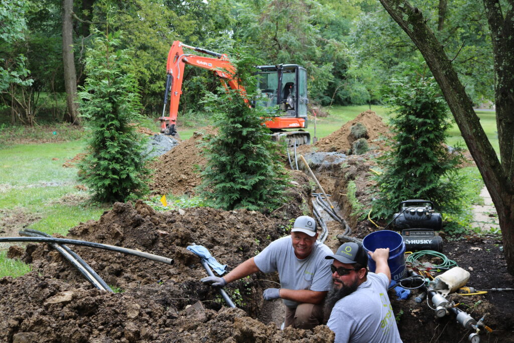Two Arronco plumbers in a ditch, working on plumbing pipes through a yard